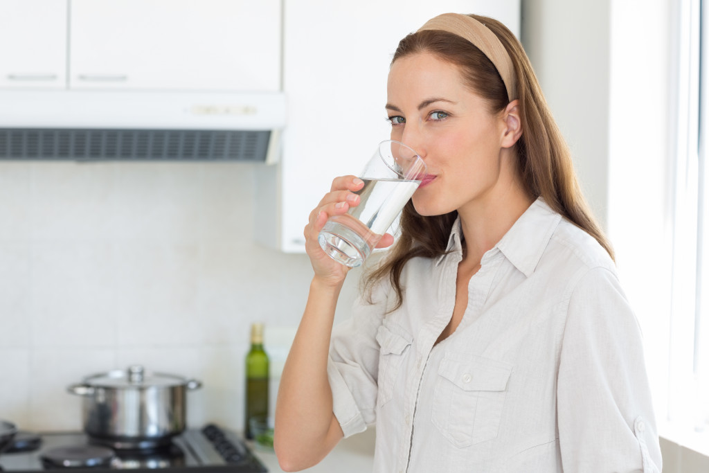 Young woman standing in kitchen and drinking water to stay hydrated 